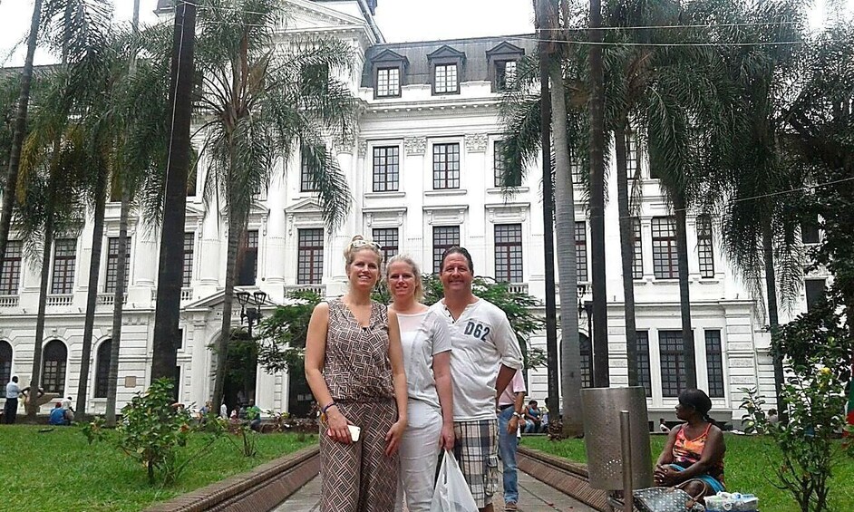 A group of people standing in front of a large white building with columns and a garden with palm trees. 4