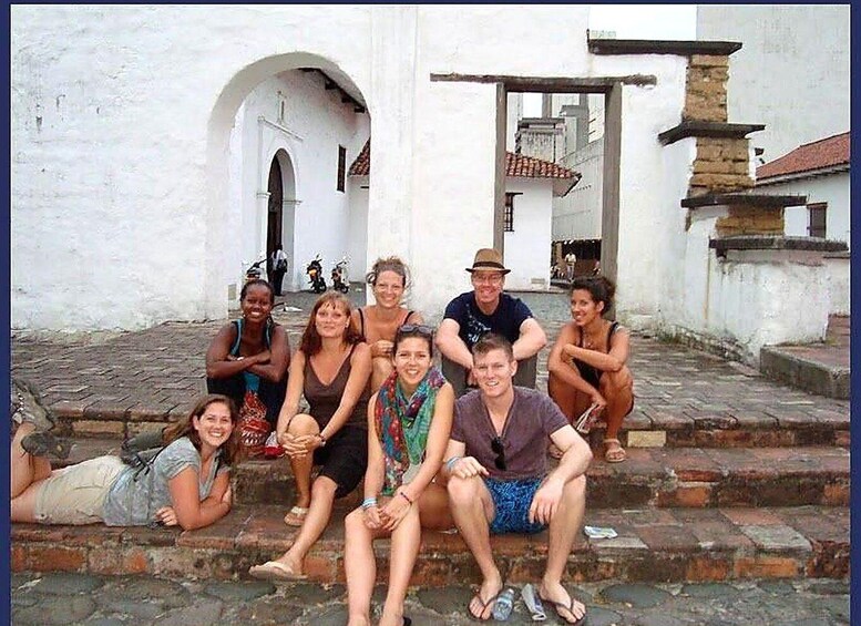 A group of people sitting on stone steps in front of a white building with an arched doorway and a tiled roof. 5