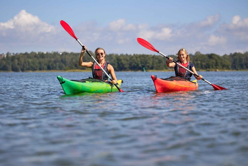 Two people kayaking on a calm lake with trees in the background. 1