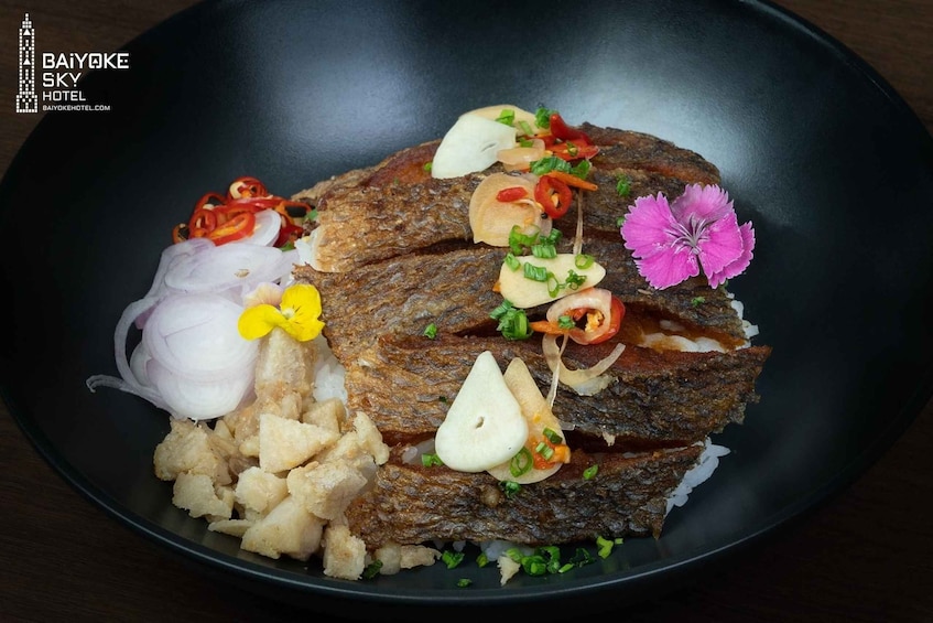 A black plate with a grilled fish topped with garlic, red chili, and a pink flower, accompanied by white rice and fried potato pieces. 2