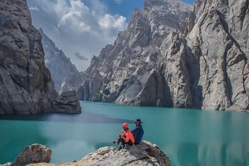 Eine gebirgige Landschaft mit einem türkisblauen See und zwei Personen, die auf einem Felsen sitzen. 3