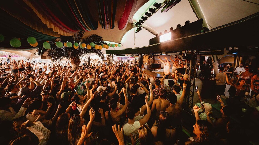 A crowded indoor event with people raising their hands, colorful decorations, and a stage with lighting equipment. 2