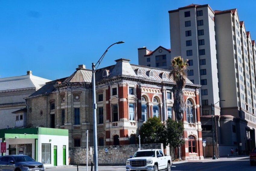 Un edificio histórico con fachada roja, un auto blanco estacionado al frente y un alto edificio moderno al fondo. 5