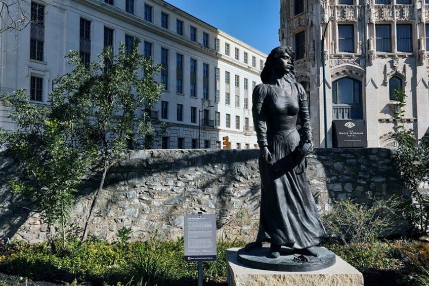 Una estatua de una mujer con vestido largo, una pared de piedra y un edificio de arquitectura clásica al fondo. 4