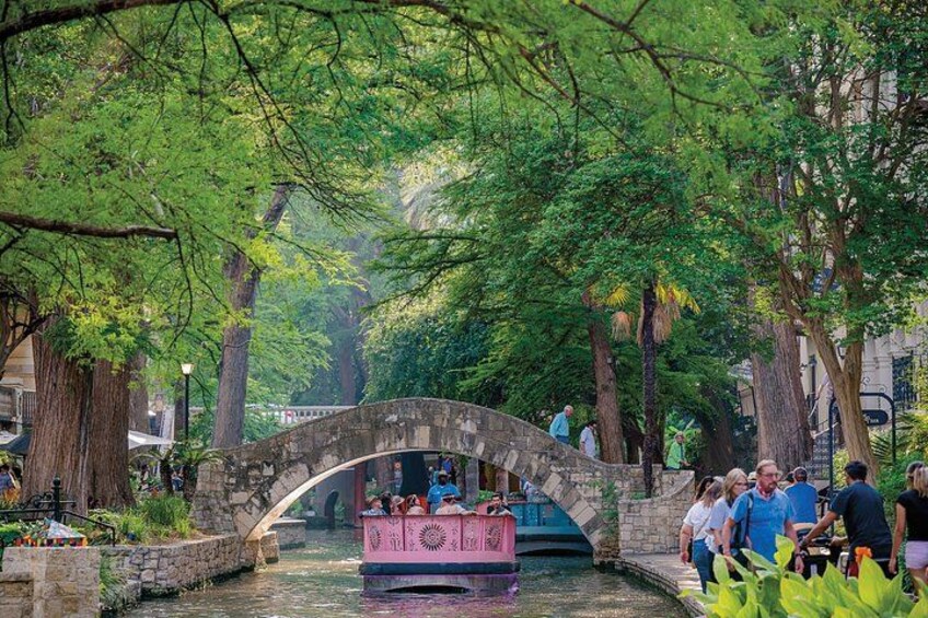 A canal with a pink boat, a stone bridge, and people walking around. 3