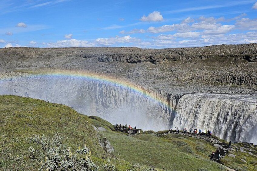 A waterfall with a rainbow, surrounded by rocky terrain and a small group of people. 1