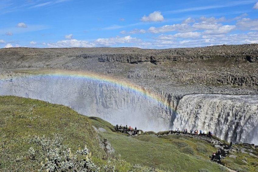 A waterfall with a rainbow, surrounded by rocky terrain and sparse vegetation. 1