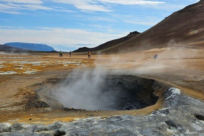 A geothermal area with steam vents, a crater, and a few people observing. 3