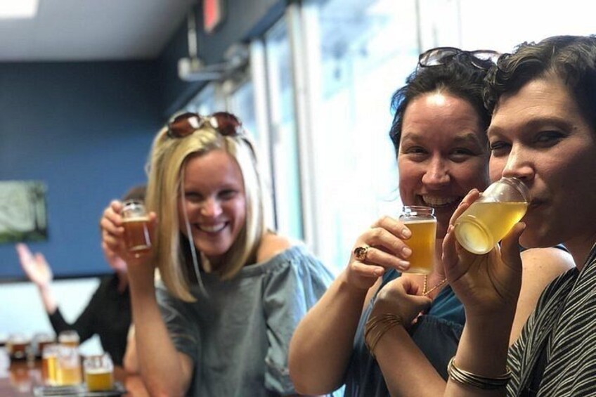 Three people holding glasses of a yellowish beverage, likely beer, in a casual indoor setting with a window in the background. 1