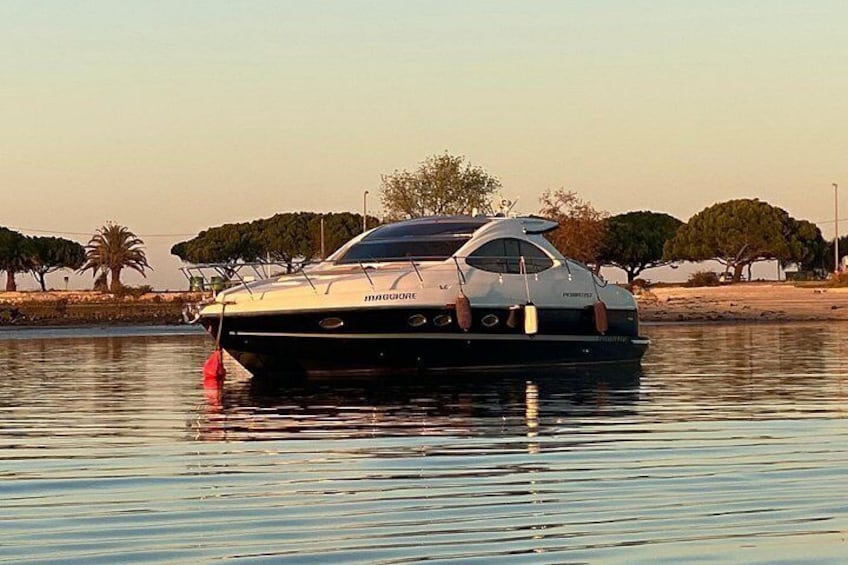 Een boot genaamd 'Maggiorino' afgemeerd in kalm water, met bomen en een heldere lucht op de achtergrond. 1