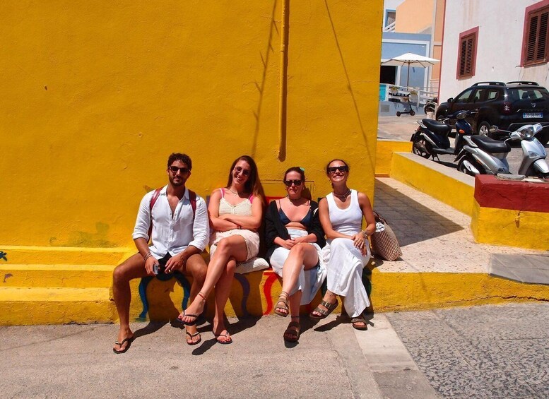 Four people sitting on a bench against a yellow wall, with a street and parked scooters in the background. 4