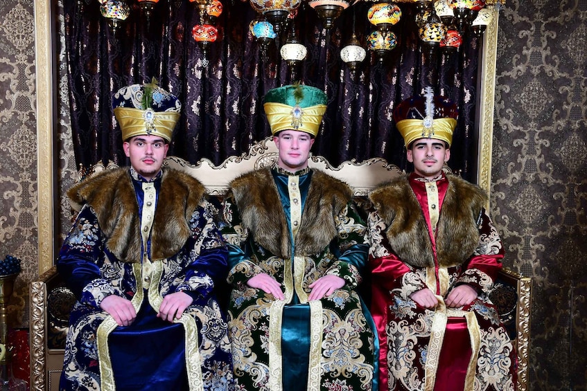 Three individuals seated on ornate thrones in a richly decorated room with patterned wallpaper and traditional, ornate headgear. 4