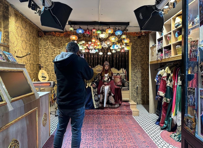 A person taking a photo of a person in traditional attire seated on a throne-like chair, surrounded by ornate lamps and patterned carpets. 2