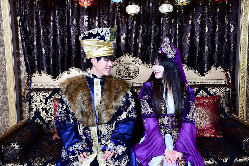 A man and woman in traditional attire seated on an ornate sofa with a patterned backdrop. 1