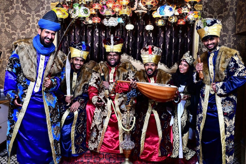 A group of people in elaborate traditional costumes with ornate headpieces and jewelry, standing in front of a richly decorated backdrop with hanging lamps. 5