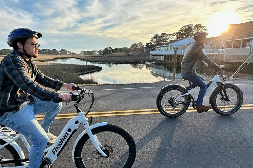 Two people riding electric bikes on a road with a body of water and a house in the background. 2