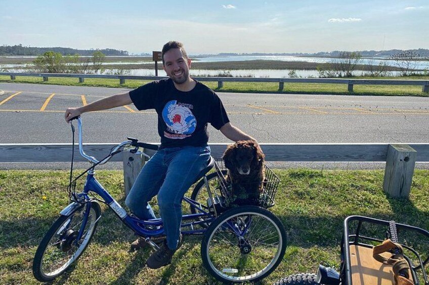 A person sitting on a bicycle with a dog in a basket, by a body of water with a wooden fence in the background. 3
