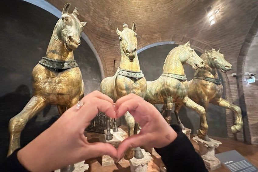 A person making a heart shape with their hands in front of golden horse statues. 2