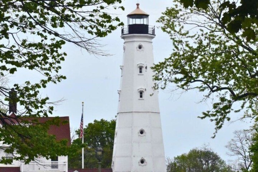 A white lighthouse with a black top, surrounded by trees and a building with a red roof. 2