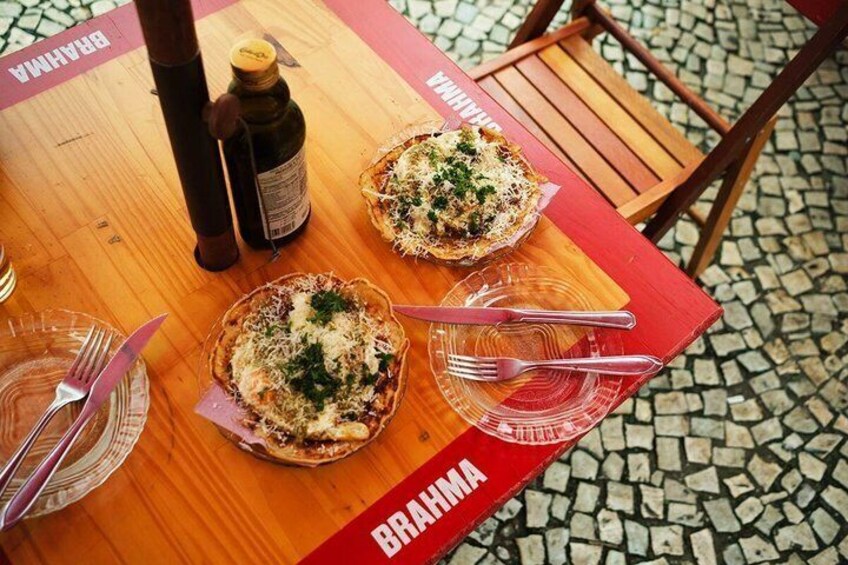 Una mesa con dos pizzas, una botella y cubiertos sobre un mantel rojo con la palabra 'BRAHMA' impresa. 3