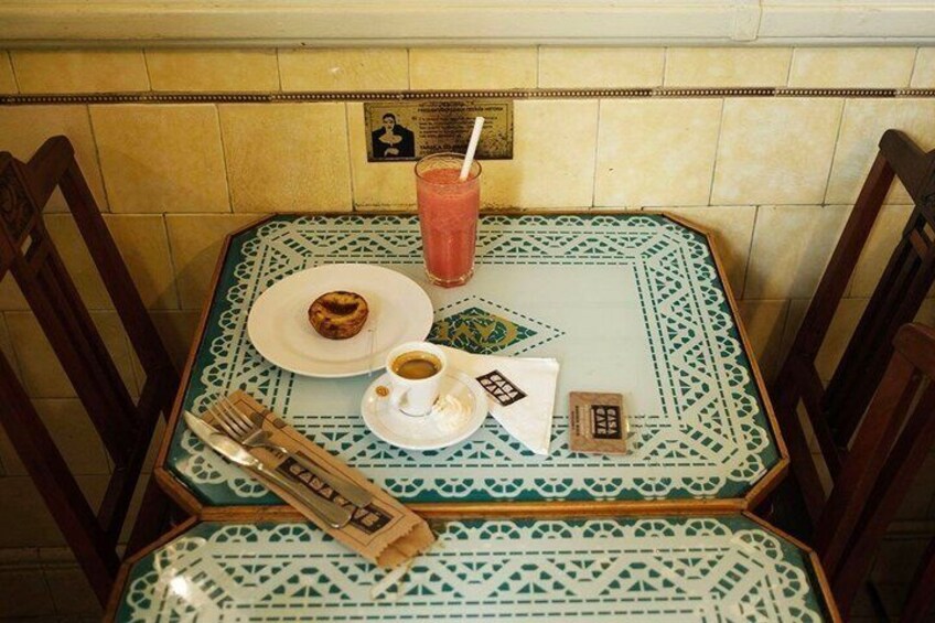 Una mesa con mantel estampado, un plato con un donut, un vaso de bebida rosada, una taza de café y un menú. 2