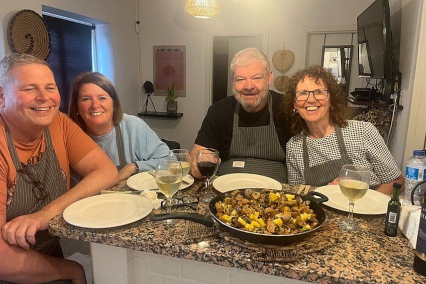 Four people seated at a kitchen counter with a pan of food, wine glasses, and a bottle of water. 3