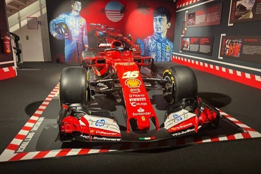 Une voiture de Formule 1 rouge, ornée des logos de ses sponsors, est exposée dans un musée, avec des panneaux d’information et un grand portrait en arrière-plan. 2