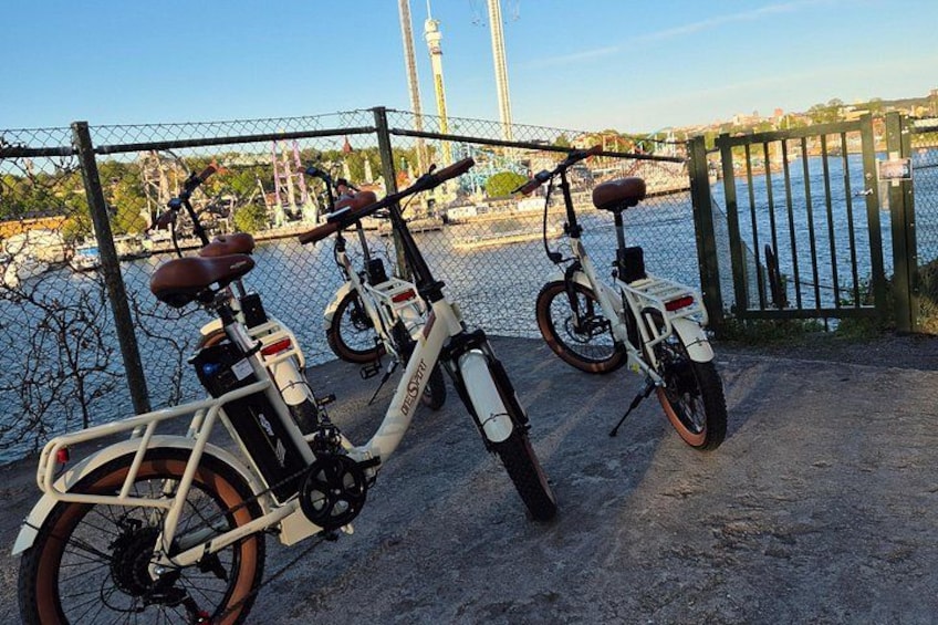 Tres bicicletas eléctricas estacionadas cerca de un muelle, con una cerca y un puente al fondo. 2