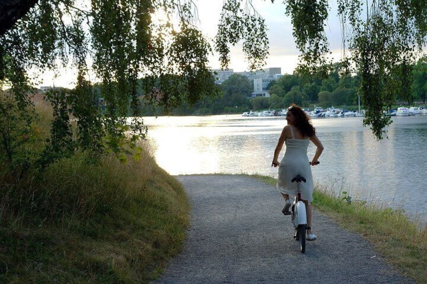 Una donna in bicicletta su un sentiero sterrato, con un lago e barche sullo sfondo. 2