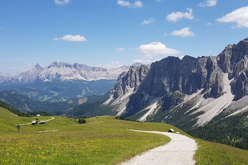 A mountain landscape with a gravel path, green fields, and rocky mountains in the background. 1