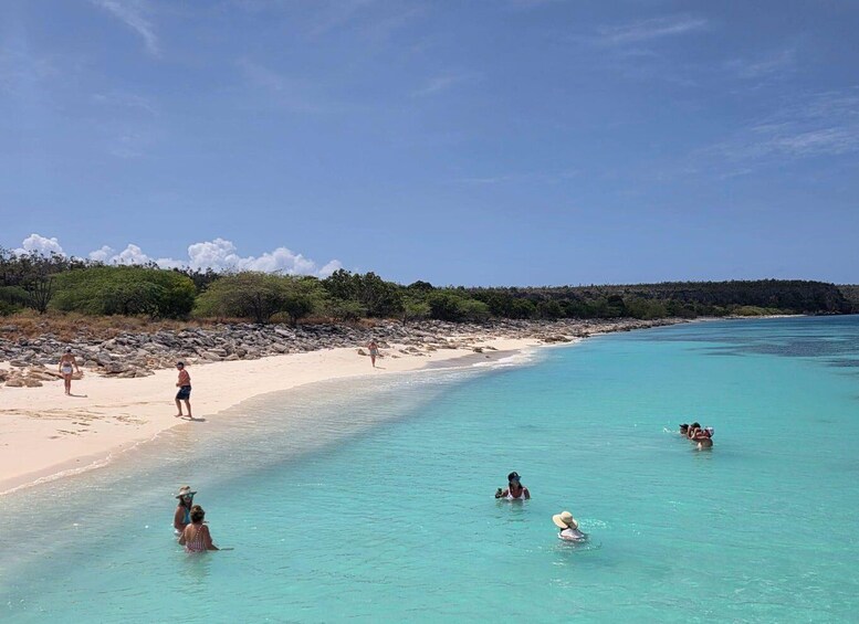 A beach with clear turquoise water, people swimming and walking on the sand, and a forested coastline. 2