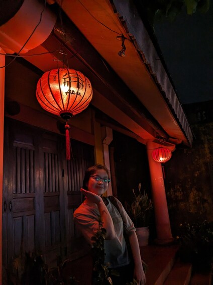 A person standing under a traditional red lantern on a porch. 1