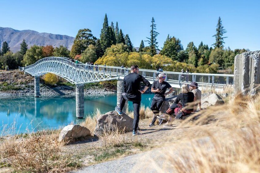 A metal bridge over a river, people walking and sitting on a path, and a mountainous landscape with trees. 1