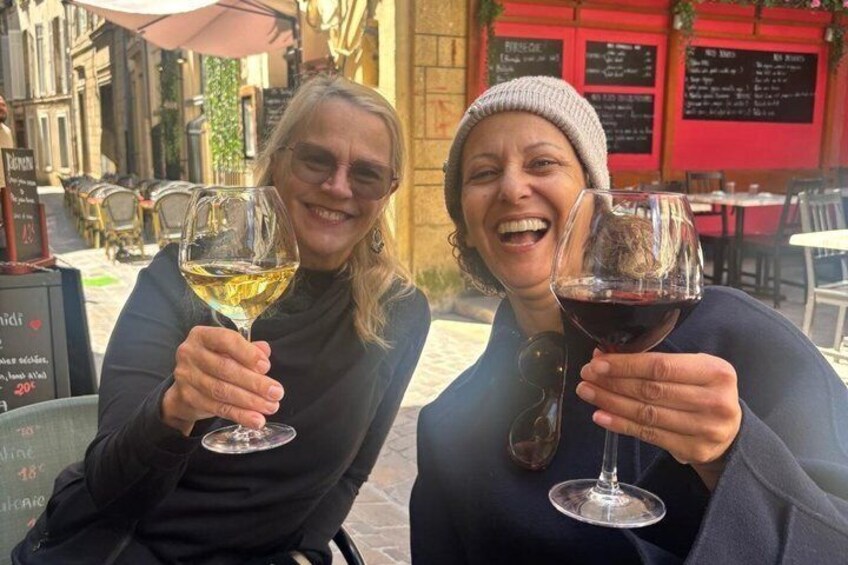 Two people holding wine glasses at an outdoor cafe with tables and chairs, and a menu board in the background. 1
