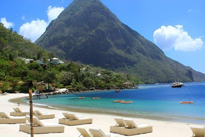 A tropical beach with clear turquoise water, lounge chairs, a mountain, and boats. 1