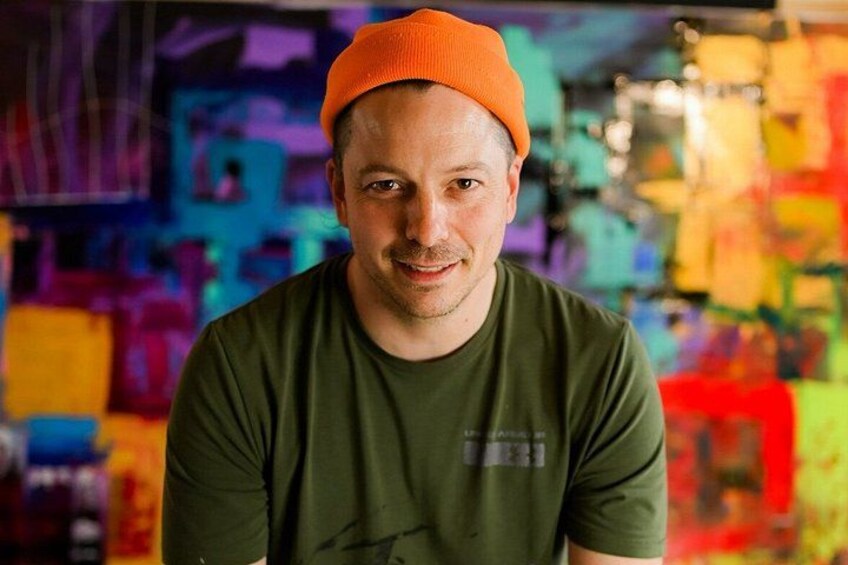 A person wearing a green t-shirt and an orange beanie, standing in front of a colorful abstract painting. 1