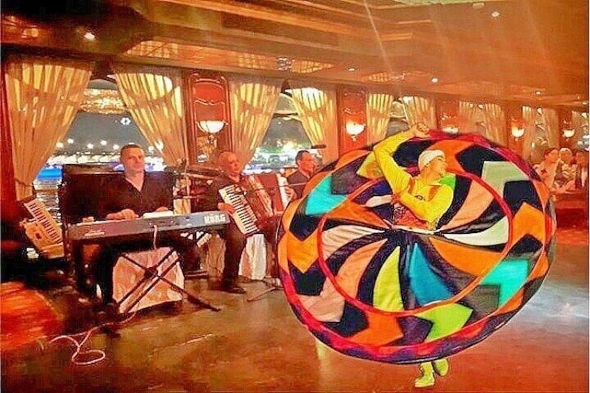 A performer in a colorful circular costume, a keyboard, and a keyboard player in a room with curtains and a cityscape in the background. 1