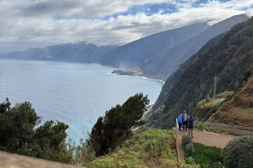 A coastal landscape with a paved walkway, a group of people, and a view of the ocean and distant mountains. 2