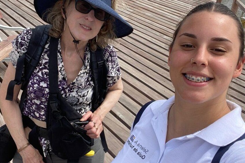Deux personnes se trouvent sur une terrasse en bois. L’une porte un chapeau et un sac à dos, l’autre est vêtue d’une chemise blanche avec un motif imprimé. 2