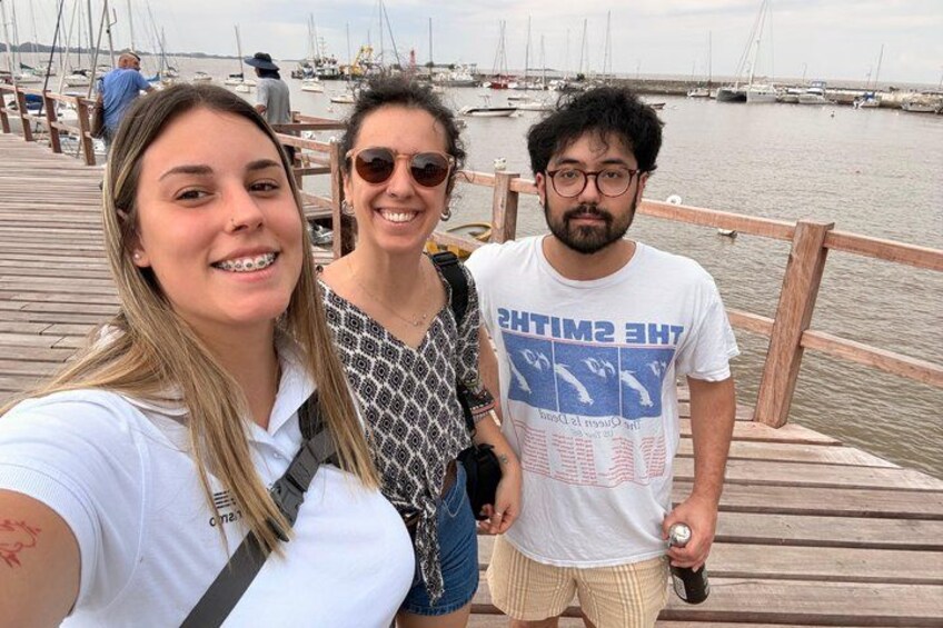 Trois personnes se tiennent sur une jetée en bois, avec des bateaux en arrière-plan. 3