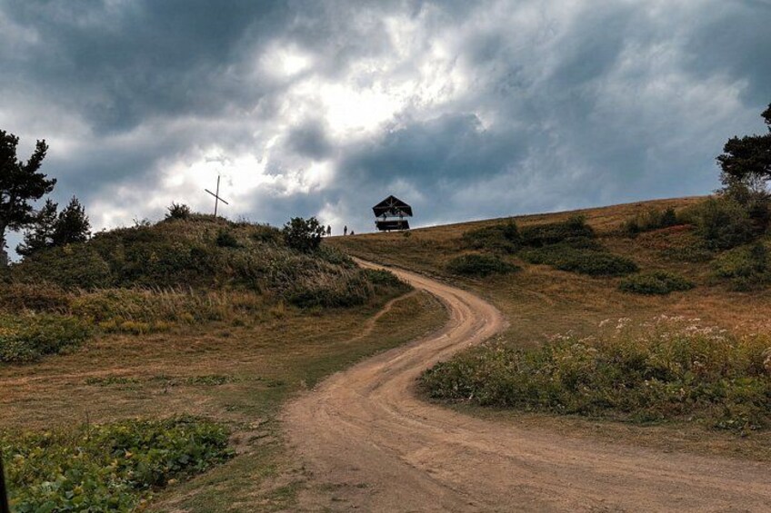 Ein Feldweg schlängelt sich durch eine hügelige Landschaft, mit einem kleinen Gebäude auf einem Hügel und einem Kreuz in der Ferne. 2
