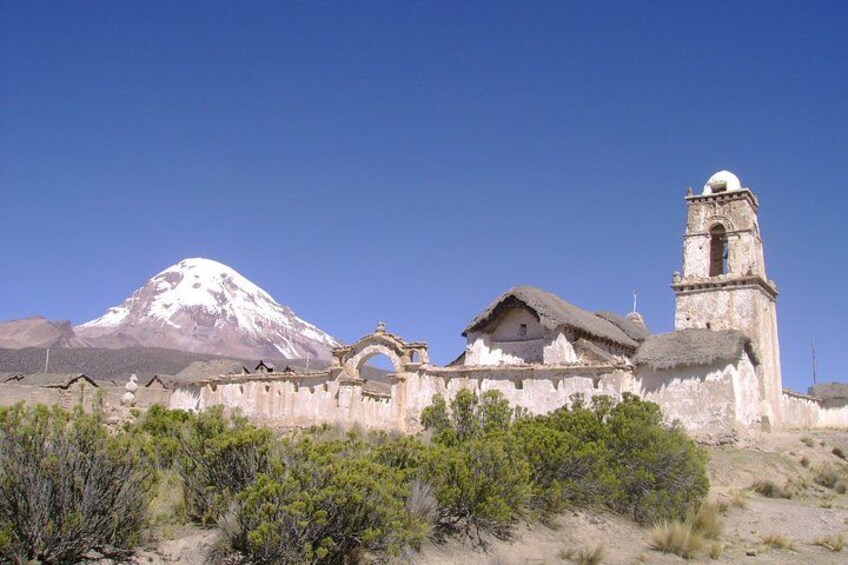 A historic church with a bell tower, a snow-capped mountain in the background, and sparse vegetation in the foreground. 3