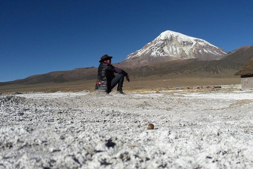 A person in a black jacket and hat sitting on a gravel surface with a snow-capped mountain in the background. 1