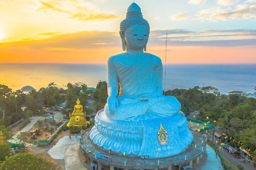 Una gran estatua de Buda con base dorada, rodeada de árboles y vista al océano al atardecer. 3
