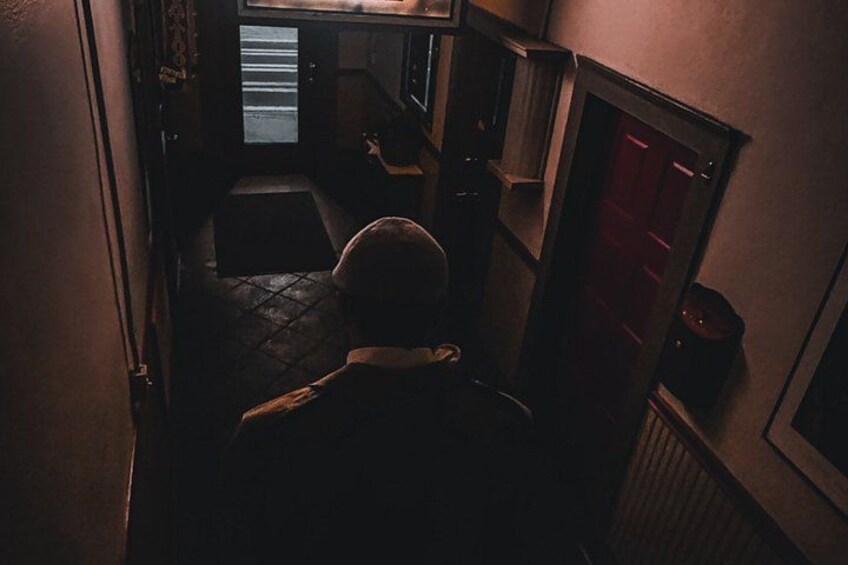 A dimly lit hallway with a person walking away from the camera, a door with a red handle, and a window with blinds. 1