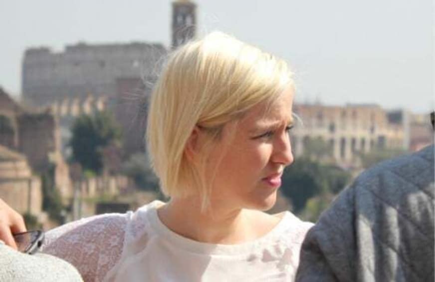 A person with blonde hair wearing a white top, standing outdoors with a historic building in the background. 4