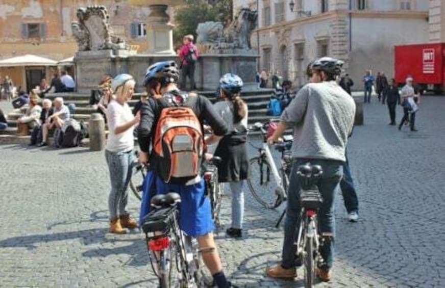 A group of cyclists in a cobblestone square with historic architecture and statues in the background. 3
