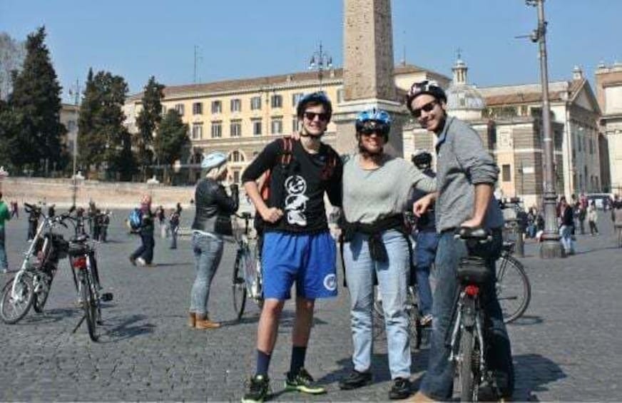 Three people standing with bicycles in a public square with historical buildings in the background. 2