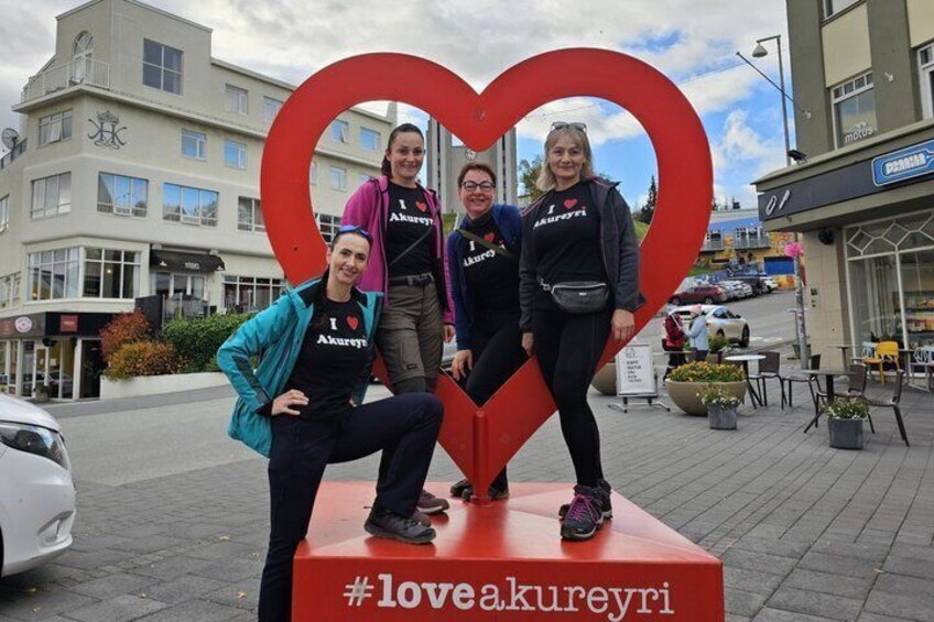 Four people posing with a heart-shaped sculpture on a pedestal, wearing matching t-shirts with a heart symbol and text. 5
