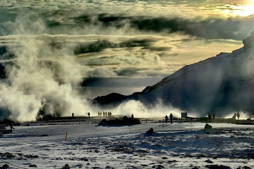 A geothermal area with steam vents, a rocky landscape, and people observing the natural features. 2